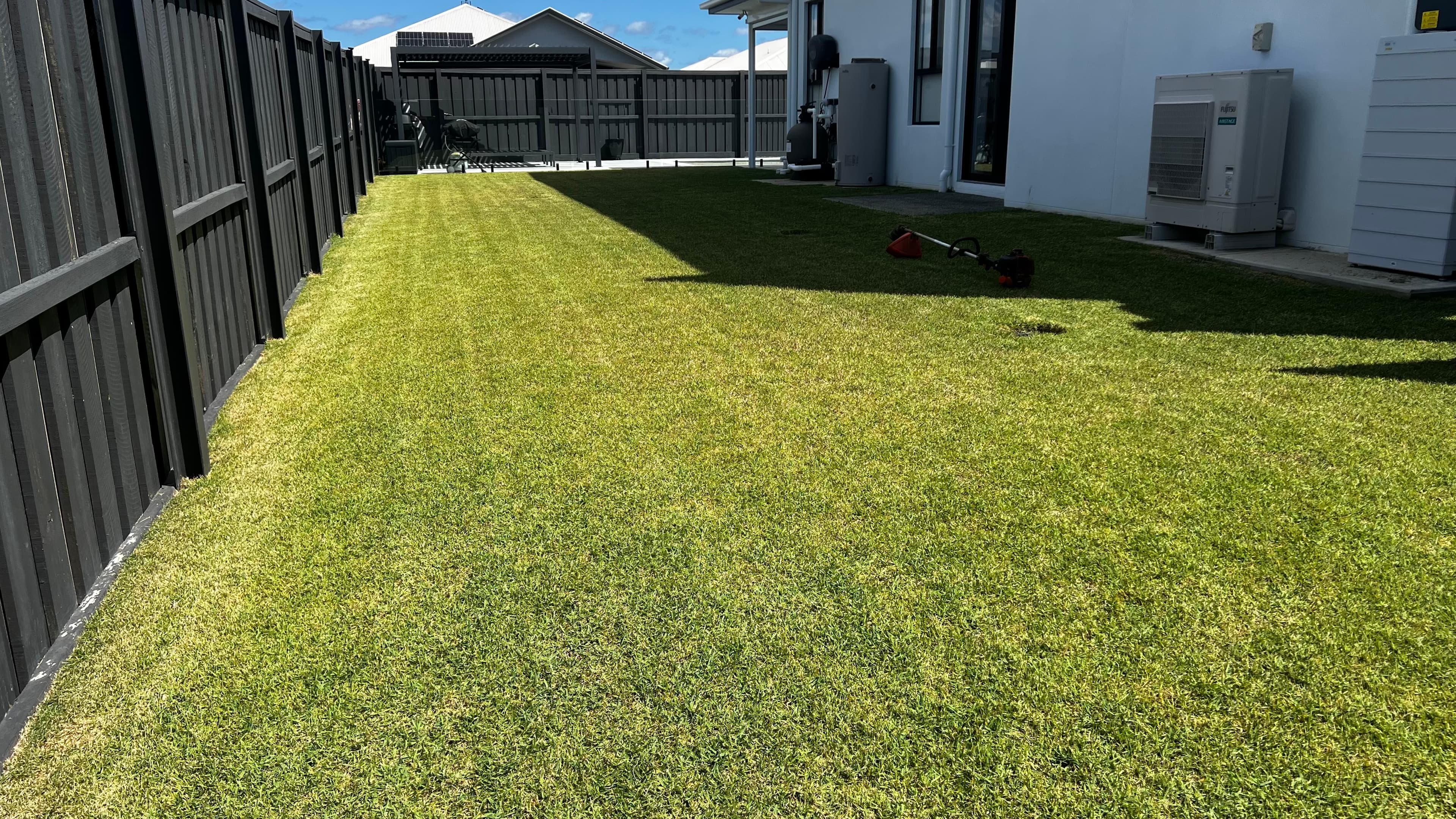 Lush green backyard lawn with a string trimmer beside a dark fence and white house.