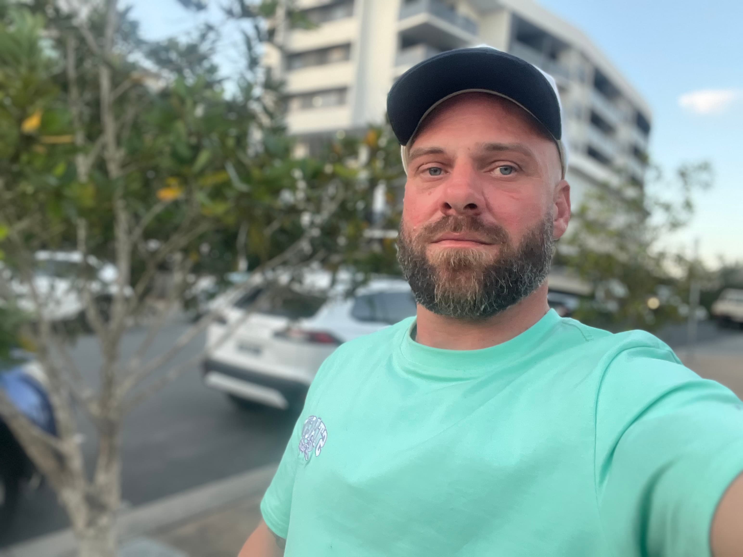 Bearded man in a mint green t-shirt and baseball cap taking a selfie outdoors.